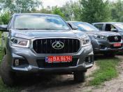 На передньому плані зображено сірий позашляховик-пікап із великою радіаторною решіткою та червоним транзитним номером «02 BA 8614», за яким на ґрунтовій дорозі на тлі дерев стоять ще два аналогічні автомобілі.