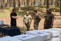 У сосновому лісі п'ятеро чоловіків у цивільному та військовому одязі стоять біля столу з розкладеними дронами та обговорюють щось поруч із великими стосами білих коробок, на яких нанесено фірмовий логотип із черешнею.