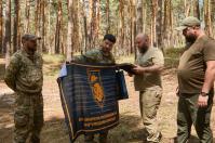 У сосновому лісі четверо чоловіків у військовій та цивільній формі розглядають і розгортають великий чорний прапор із помаранчевим написом «60 ОКРЕМА МЕХАНІЗОВАНА ІНГУЛЕЦЬКА БРИГАДА» та емблемою ведмедя по центру.