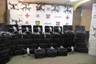 У закритому приміщенні на тлі білого банера із гербами та написом «100 ОКРЕМА МЕХАНІЗОВАНА БРИГАДА» виставлено велику кількість військової допомоги, що складається зі стосів чорних кейсів із розкладеними квадрокоптерами, а також FPV-дронів, закріплених на стіні та на коробках з обох боків.