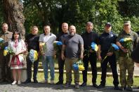 Шеренга з дев'яти людей у цивільному, військовому та поліцейському вбранні стоїть просто неба біля великого дерева, тримаючи в руках патріотичні синьо-жовті букети квітів під час офіційного заходу вшанування пам'яті на тлі зелених насаджень.