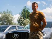 На передньому плані праворуч стоїть чоловік у коричневій футболці з написом «ARMY OF UKRAINE» та камуфляжних штанах, тримаючи руки за спиною, а на задньому плані видно частину сріблястого позашляховика, дерева та блакитне небо з білими хмарами.