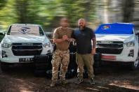 На ґрунтовій дорозі між двома білими пікапами Isuzu з прапорами на лобовому склі стоять двоє чоловіків — ліворуч військовий у камуфляжних штанах із замиленим обличчям, а праворуч бородатий чоловік у чорній футболці, які міцно тиснуть один одному руки на тлі складених на землі чорних кейсів із технікою.