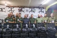 У закритому приміщенні дев'ятеро чоловіків у військовій та цивільній формі стоять у ряд, демонструючи невеликі посвідчення, перед стіною з розвішаними FPV-дронами та банером «100 ОКРЕМА МЕХАНІЗОВАНА БРИГАДА», а перед ними на підлозі виставлено два ряди великих чорних кейсів із розпакованими квадрокоптерами.