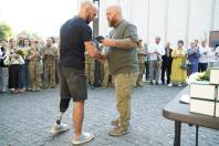 Лисий чоловік у зеленій футболці тисне руку та вручає нагороду ветерану з протезом лівої ноги, який одягнений у чорну футболку та шорти, на брукованій площі перед групою військових та цивільних людей.