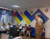 На світлині в робочому кабінеті сивий чоловік у військовій формі в профіль спілкується з групою цивільних людей, які сидять на стільцях та синіх кріслах на тлі стіни з трьома великими прапорами, серед яких Державний Прапор України.