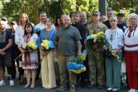 Великий натовп людей, серед яких є військовослужбовці, поліцейські та цивільні у вишиванках, зосереджено стоїть на вулиці біля дерев, тримаючи в руках великі синьо-жовті букети квітів під час офіційного заходу.