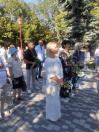Група людей у вишиванках та літньому вбранні зосереджено стоїть на брукованій парковій алеї під час офіційного заходу, тримаючи в руках букети квітів поруч із великою ялиною та зеленими деревами.