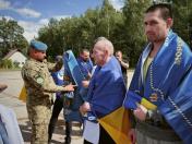 На світлині просто неба військовослужбовець у камуфляжі та блакитному береті огортає синьо-жовтими державними і військовими прапорами звільнених із полону чоловіків, один з яких на передньому плані дивиться в об'єктив.