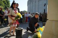 Чоловік у чорному одязі опустився на одне коліно, щоб покласти синьо-жовті квіти до підніжжя пам'ятника, біля якого стоїть жінка у вишиванці з букетом хризантем та група людей на брукованій площі під час урочистої церемонії.