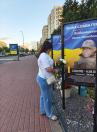 Жінка у білій футболці та синіх джинсах стоїть на брукованому тротуарі біля ряду меморіальних стендів із синьо-жовтим тлом і портретами полеглих бійців, поправляючи білі троянди біля плаката з написом «Вічна слава героям».