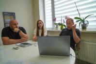 У кабінеті біля вікна з орхідеями троє людей — двоє чоловіків та дівчина — сидять за круглим білим столом і зосереджено дивляться на екран ноутбука перед ними.