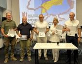 П'ять учасників заходу, серед яких військовий та жінка у світлому костюмі, стоять у ряд за білим столом та демонструють у руках підписані ювілейні поштові конверти на тлі великого екрана з картою України.