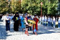 Урочиста хода рухається брукованою алеєю парку, де попереду дівчина та хлопець у яскравих традиційних українських костюмах несуть великий кошик із синьо-жовтими квітами, а за ними йдуть священник у чорній рясі та група людей у цивільному одязі й вишиванках.