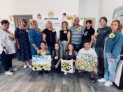 У світлій кімнаті група дорослих та підлітків позує для спільного фото, тримаючи в руках картини та вироби із соняшниками, при цьому одяг багатьох учасників також прикрашений жовтими квітками соняшника.