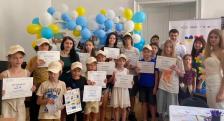 У світлій кімнаті велика група дітей та дорослих позитивно позує для спільного фото, тримаючи в руках отримані сертифікати на тлі великої фотозони з білих, жовтих і блакитних повітряних кульок.