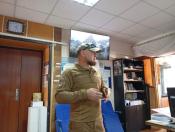 На світлині в робочому кабінеті лисий чоловік із рудою бородою у військовій формі та кепці стоїть по центру та жестикулює руками, а на задньому плані видно великий настінний екран із гірським пейзажем, сині крісла та шафу з книгами.