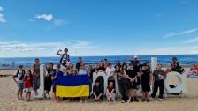 Велика група дітей та дорослих позирує для спільного фото на піщаному пляжі біля моря, тримаючи розгорнутий синьо-жовтий прапор України перед великим білим написом «Jūrmala»