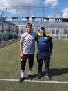 Двоє чоловіків у спортивній формі стоять обнявшись за плечі на футбольному полі зі штучним покриттям на тлі високого паркану, трибун та хмарного неба.