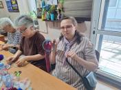 На зображенні жінка в окулярах та смугастій сукні сидить за довгим столом поруч із двома іншими учасницями та показує в руці незавершений ловець снів із блакитним обручем і рожевими нитками, поки інші жінки продовжують роботу над своїми виробами.