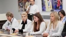 На зображенні у приміщенні за довгим темним столом сидять чотири жінки та слухають виступ чоловіка у білій футболці, який стоїть на задньому плані з мікрофоном у руках на тлі стіни з великими фотографіями спортсменів.