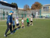 На футбольному полі зі штучним покриттям тренер у синьо-жовтій спортивній кофті та синій кепці веде за собою вервечкою п'ятьох маленьких дітей, які тримаються за руки, на тлі високої огорожі, трибун та дерев.