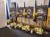 На зображенні у приміщенні біля бетонної стіни в ряд на дерев'яних мольбертах встановлено чотири траурні плакати з чорно-білими портретами й іменами загиблих українських військових та журналістів — Олександра Махова, Миколи Сіренка, Дмитра Сірика та Андрія Ширшикова, біля кожного з яких стоять жовто-блакитні прапори України, кошики з жовто-білими квітами та запалені лампадки.