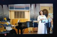 Скриншот відеотрансляції виступу в залі з великим органом, де на сцені біля чорного рояля C. Bechstein стоїть співачка в білій сукні з вишивкою перед закріпленим на штативі смартфоном, ліворуч за інструментом сидить піаністка, а поверх відео відображаються багаторазові накладення імені глядача «Любов Сидорова» та смайлики-оплески.