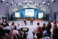 Двоє чоловіків у костюмах сидять у кріслах на круглій сцені та ведуть дискусію перед аудиторією під час саміту «CARPATHIAN», на тлі великих мультимедійних екранів із гірськими пейзажами та професійного освітлення.