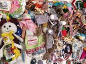 На зображенні представлена велика різнобарвна колекція вироблених вручну сувенірів та оберегів, серед яких виділяються традиційні українські ляльки-мотанки в етнічному вбранні з вишивкою, велика м'яка іграшка ведмедика з бантом та численні плетені прикраси й браслети.