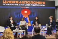 На зображенні четверо спікерів — один чоловік та три жінки — сидять на стільцях на сцені під час панельної дискусії на тлі великого темного екрана з помаранчево-білим написом «ЦИВІЛЬНІ ПОЛОНЕНІ: ПОВЕРНУТИ, ВИЗНАТИ, ПІДТРИМАТИ» та символічним малюнком замкової щілини.