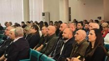У великій залі з колонами та жалюзі на вікнах на сидіннях рядами сидять численні слухачі, які зосереджено дивляться в один бік.