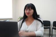 У світлому офісному кабінеті за столом перед ноутбуком сидить брюнетка з чубчиком у білій кофті, зосереджено дивлячись на екран.