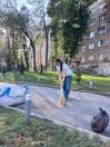 Жінка у білій футболці та джинсах замітає віником асфальтовану доріжку біля скверу та багатоквартирного будинку, де на узбіччі лежать наповнені мішки для сміття.