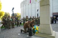 На світлині двоє українських військовослужбовців у тактичних куртках приклонили коліно, покладаючи великий жовто-блакитний букет квітів до підніжжя високого світлого монумента на тлі шеренги інших побратимів у камуфляжі.