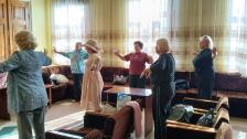 На зображенні шість жінок старшого віку займаються руховою гімнастикою в просторій світлій кімнаті з великими вікнами, стоячи біля крісел і диванів та повторюючи вправи з витягнутими або розведеними в боки руками.