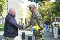На світлині бородатий чоловік у темній жилетці тисне руку українському військовослужбовцю у камуфляжній формі та кепці, який тримає букет жовто-блакитних квітів, на тлі кам'яного монумента та інших присутніх людей.