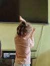 На зображенні маленька дівчинка з зібраним у пучок волоссям стоїть спиною до глядача в рожевій кофті на тлі світло-жовтої стіни та великого вимкненого телевізора, тримаючи підняті руки перед собою із зімкнутими долонями.