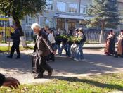 На світлині зображено урочисту процесію на асфальтованій дорозі біля будівлі, де літній чоловік несе портативну аудіоколонку, група молоді несе хвойну гірлянду-вінок, а попереду та позаду них ідуть чоловік із хоругвою та священнослужителі в рясах.