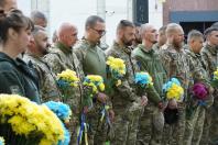 На світлині шеренга українських захисників і захисниць у камуфляжній формі стоїть із похиленими головами під час хвилини мовчання, тримаючи в руках патріотичні жовто-блакитні та фіолетові букети квітів.