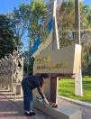 На світлині дівчина у чорній куртці схилилася, щоб покласти червоні троянди до підніжжя меморіальної стели з написом «Герої не вмирають!», увінчаної гербом та прапором України.