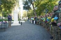 На світлині чоловік із мікрофоном виступає перед шеренгою військовослужбовців та цивільних із жовто-блакитними квітами, які зібралися на брукованій площі біля світлого кам'яного монумента серед дерев.