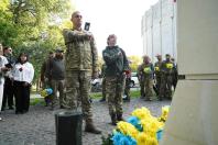 На світлині двоє українських військовослужбовців — чоловік та жінка в камуфляжній формі — стоять струнко, приклавши праву руку до серця, біля підніжжя кам'яного монумента з жовто-блакитними квітами на тлі інших присутніх людей.