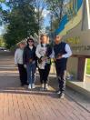На світлині четверо людей стоять на брукованій алеї біля стели з написом «Герої не вмирають», тримаючи в руках червоні троянди на знак вшанування пам'яті.