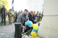 На світлині двоє чоловіків присіли, щоб покласти великі жовто-блакитні букети квітів до підніжжя світлого кам'яного монумента, на тлі інших цивільних та військових, які стоять поруч на брукованій площі.