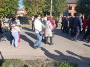 На світлині група людей різного віку йде вздовж асфальтованої вулиці повз зелені дерева та будівлі, причому деякі жінки у натовпі тримають у руках маленькі жовто-блакитні українські прапорці.