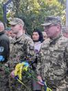 На світлині двоє українських військовослужбовців у камуфляжній формі та кепках стоять у шерензі, тримаючи в руках патріотичні жовто-блакитні квіти, а позаду них у натовпі видно цивільну жінку на тлі зелених дерев.