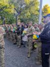 На світлині велика шеренга військовослужбовців ЗСУ, поліцейських та цивільних громадян стоїть на брукованій алеї біля парку, тримаючи в руках патріотичні жовто-блакитні букети квітів під час урочистого заходу.