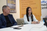 Молода жінка з довгим темним волоссям у світлій сорочці сидить за білим столом і емоційно говорить, жестикулюючи руками, у той час як чоловік у синьому піджаку ліворуч уважно її слухає на тлі інформаційних банерів