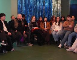  Близько п'ятнадцяти осіб різного віку (переважно молодь) зосереджено слухають або спостерігають за чимось поза кадром. Вони одягнені в повсякденний одяг: худі, джинси, куртки та светри.